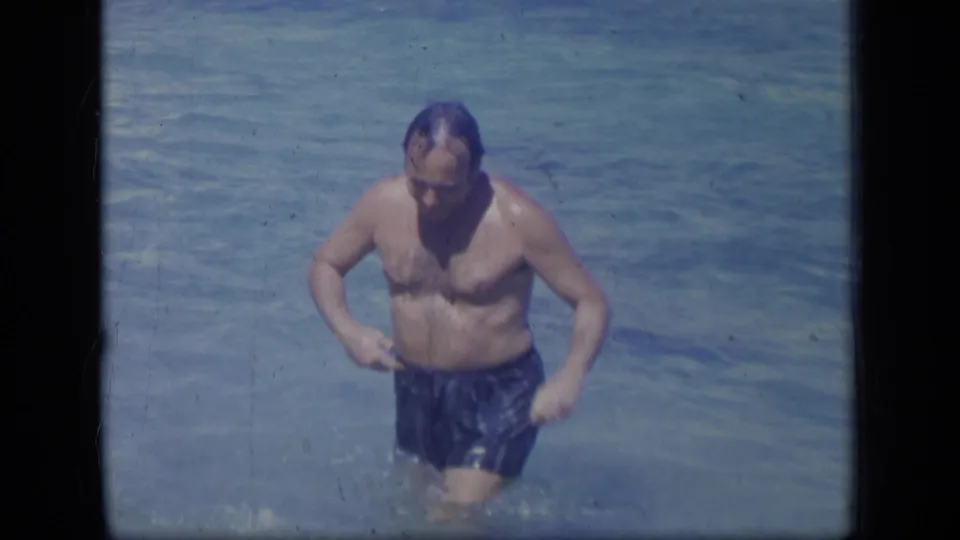 An adult man in blue swim trunks wades through clear Puerto Rico shallows, looking down as water splashes around his legs.