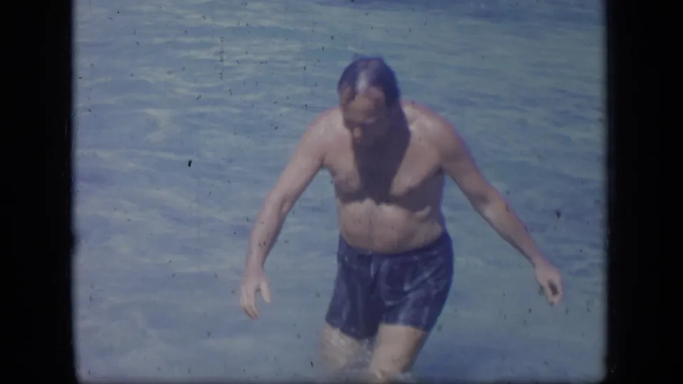 A 1970s home-movie frame shows an adult man in blue swim trunks walking forward through clear shallow water in Puerto Rico.