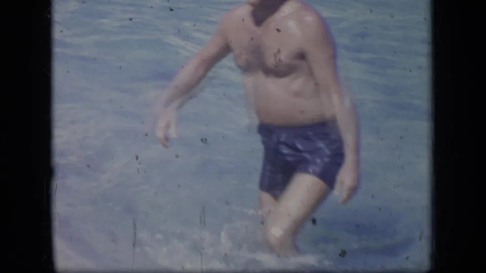 The man in blue swim trunks is partly cropped at the edge of the frame as he steps through clear shallow water.