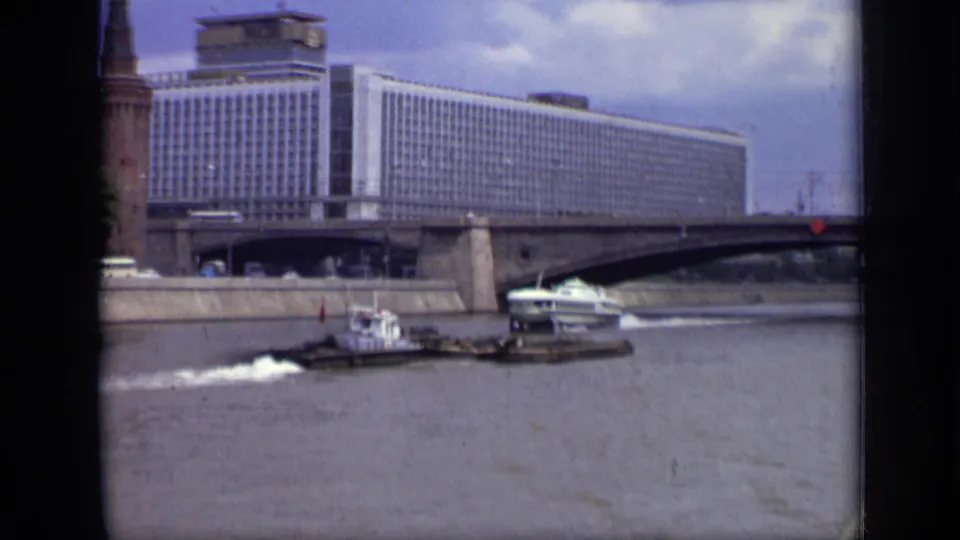 Closer Moscow riverfront frame with Hotel Ukraina behind a bridge, a tower-like structure, boat wake, and vehicles near the embankment.
