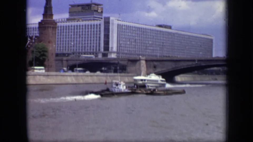 1971 Moscow river view with a white hydrofoil, tugboat, barge, bridge, Hotel Ukraina, and Kremlin tower in the background.
