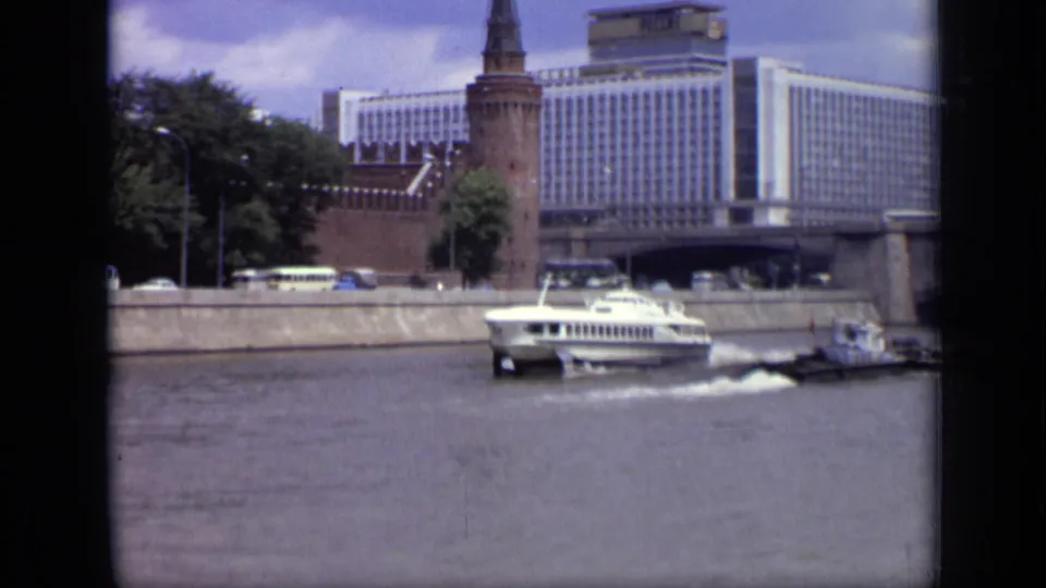 1971 Moscow waterfront frame showing a white hydrofoil moving past buses, trees, a brick tower, and Hotel Ukraina.