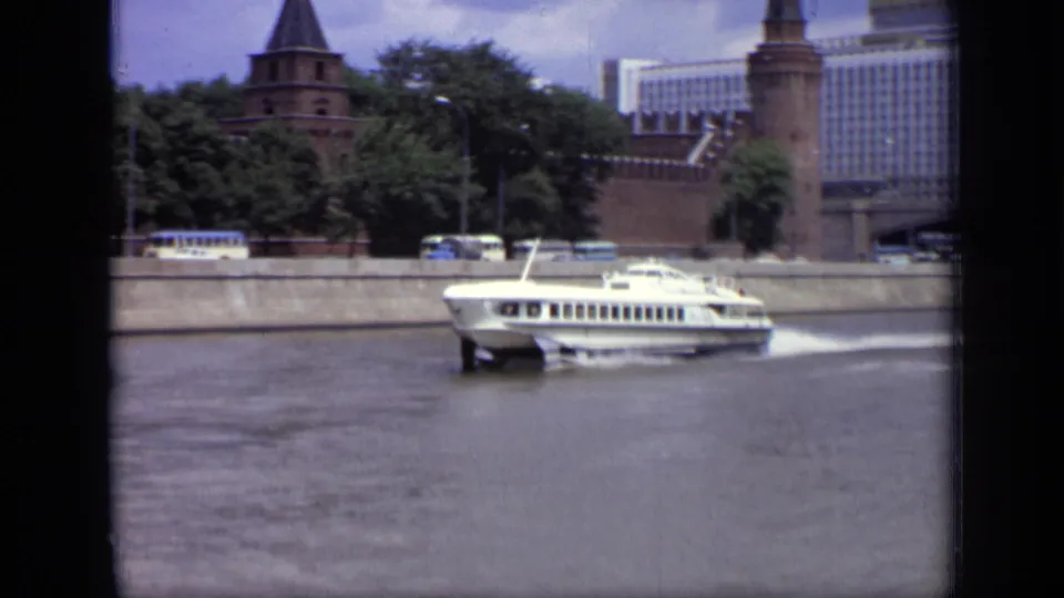 White hydrofoil crosses a Moscow river frame with embankment buses, Kremlin brick towers, trees, and Hotel Ukraina behind.
