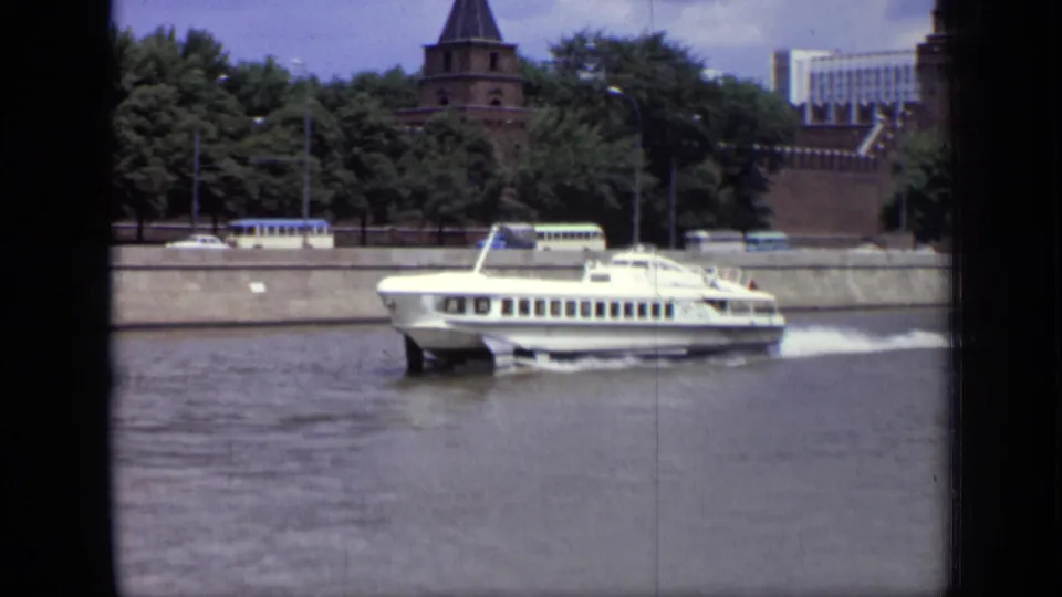 1971 Moscow 8mm frame of a white hydrofoil crossing the river, with Kremlin walls, towers, trees, and embankment traffic in the background.