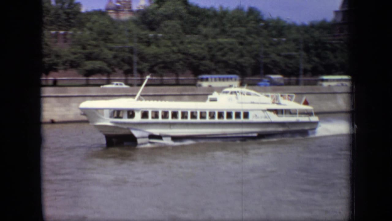 Long white hydrofoil boat passing across the river beside a tree-lined embankment.