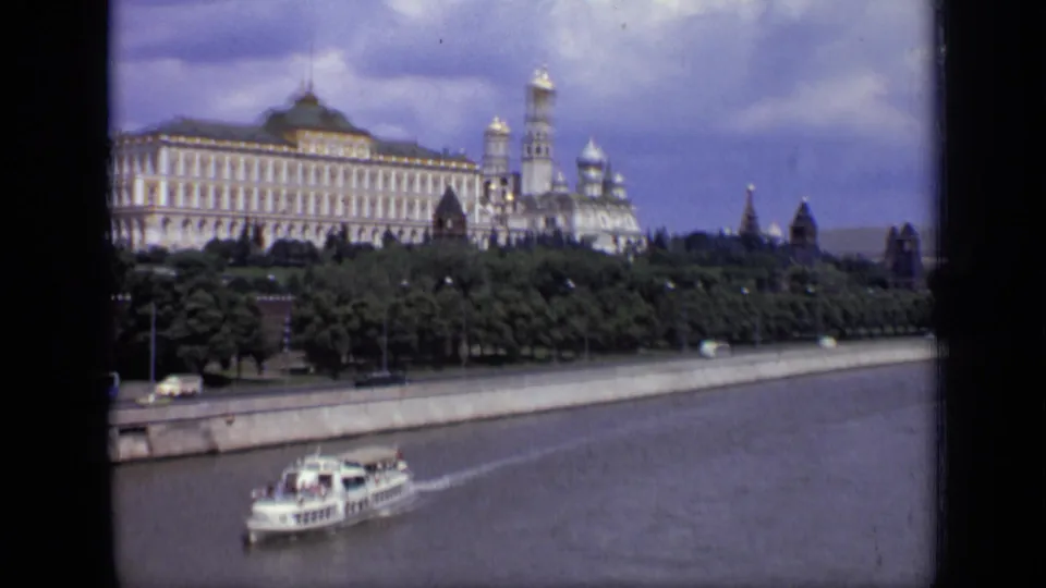 1971 Moscow 8mm frame widening to show a small river boat beneath Kremlin buildings, towers, trees, embankment, and a partly cloudy sky.