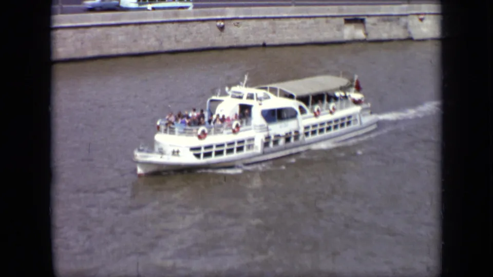 Close 1971 Moscow river view of a white excursion boat with passengers visible on deck near the embankment.