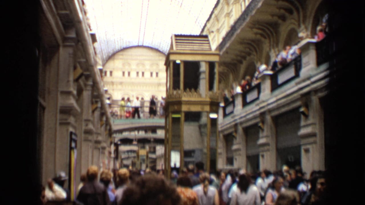 1971 Moscow home-movie frame showing a busy shopping arcade with crowds below, people on balconies, and a glass-vaulted roof.