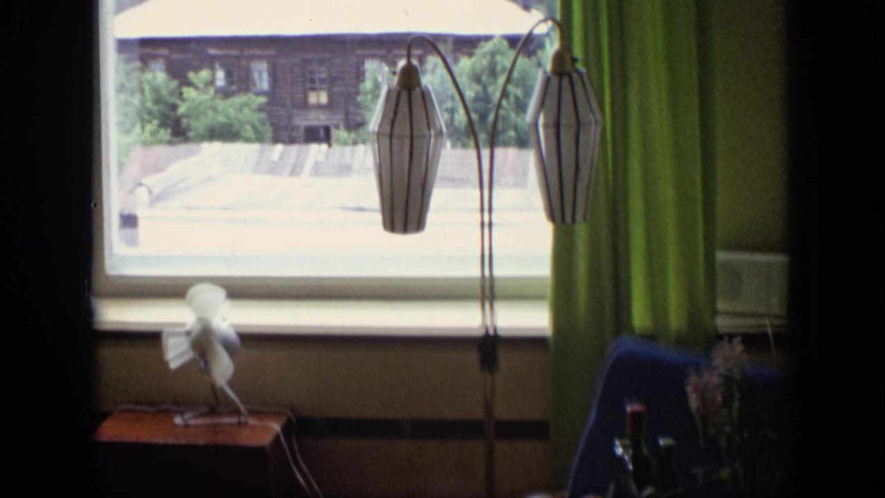Apartment window with green curtains, striped floor lamp, small fan, and a wooden house outside.
