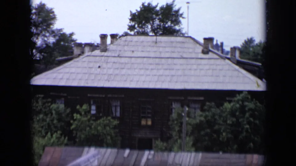 Closer 1971 Moscow window view of a wooden house with a pale roof and leafy trees partly covering the facade.