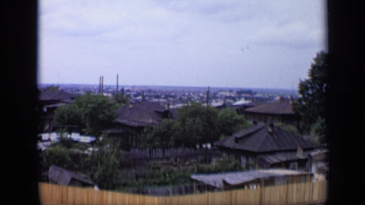 1971 Moscow overlook with low wooden rooftops, leafy trees, a fenced plot, cloudy sky, and distant industrial structures.