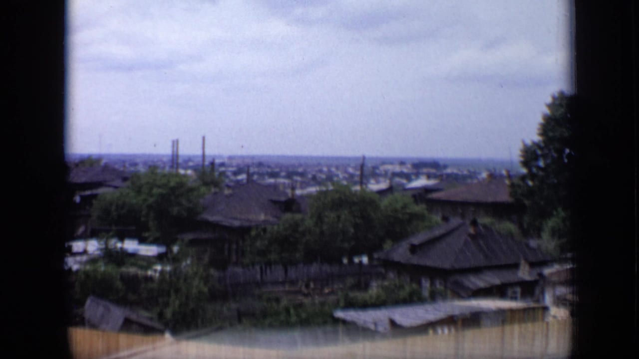 1971 Moscow rooftop view showing low residential buildings, green trees, a wooden fence and distant smokestacks beyond the neighborhood.