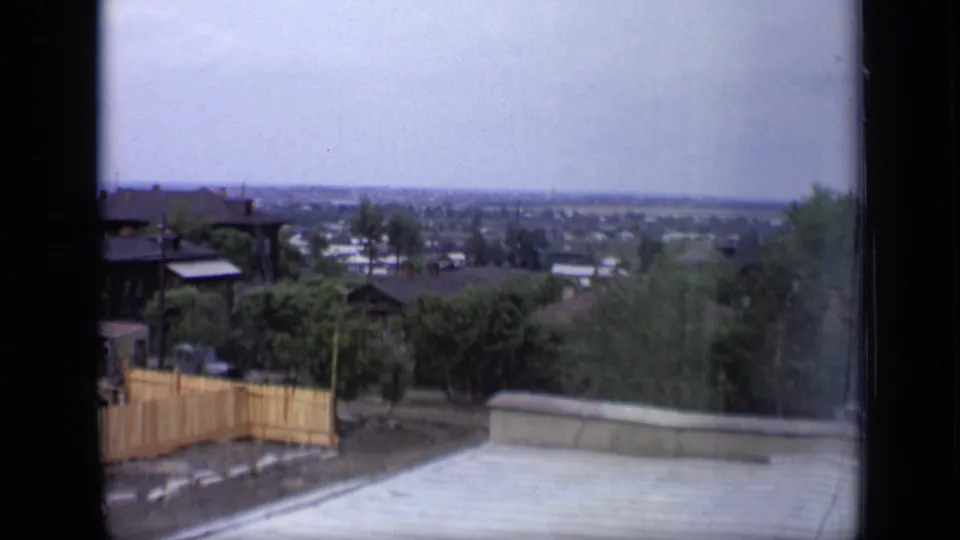 1971 Moscow view from a rooftop or terrace edge, looking past a wooden fence and trees toward a broad, hazy urban horizon.