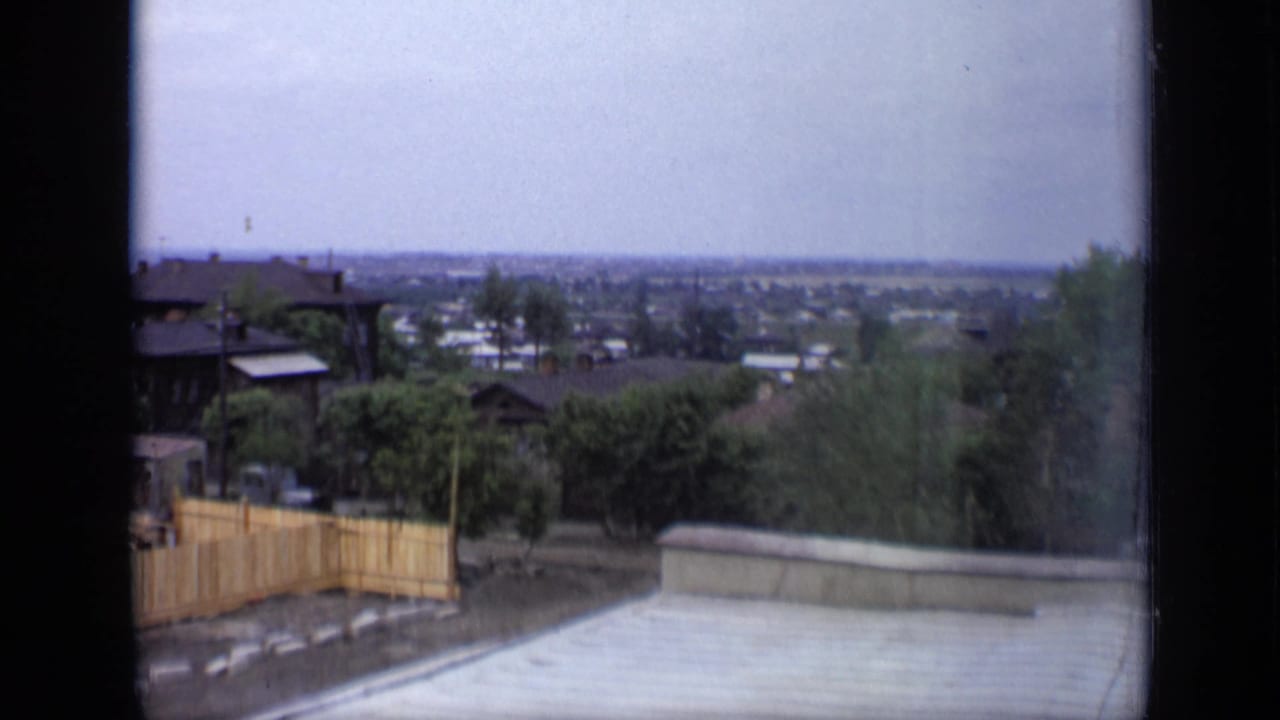 1971 Moscow home-movie view from a window toward wooden houses, greenery, fences, and a distant city skyline.