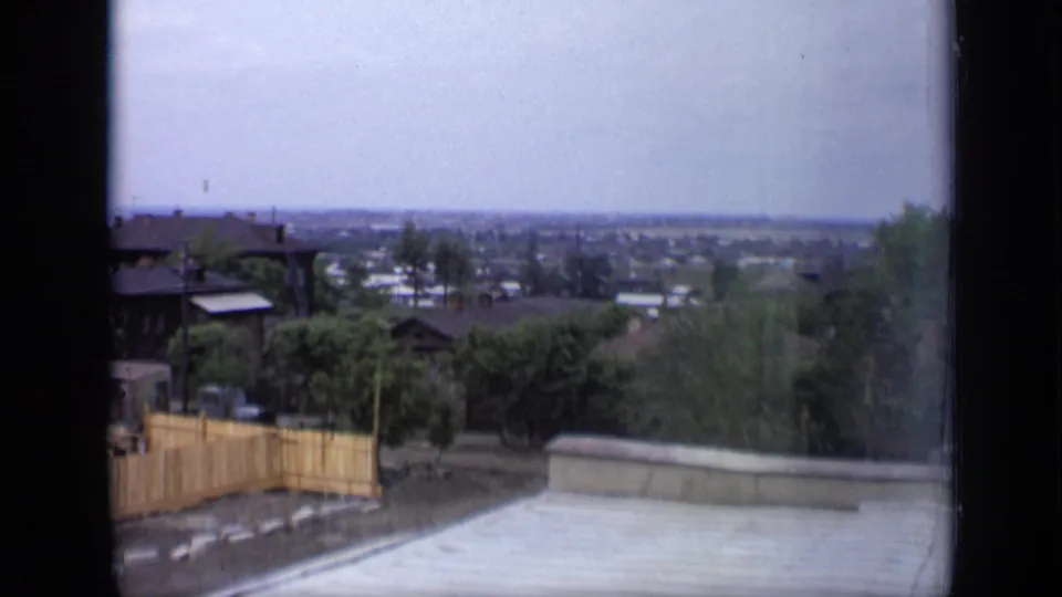 1971 Moscow home-movie view from a window toward wooden houses, greenery, fences, and a distant city skyline.