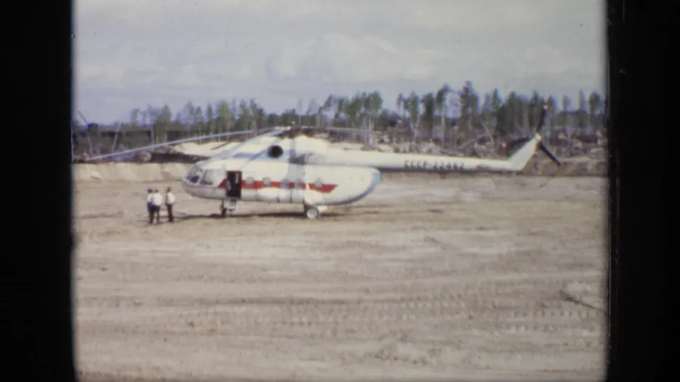 1971 Moscow 8mm frame of a white and red helicopter parked on a dirt airfield with two figures near the open door.