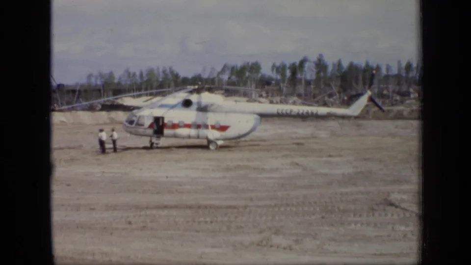 1971 Moscow home-movie frame showing a white and red helicopter on an open dirt field, with two small figures near the side door.