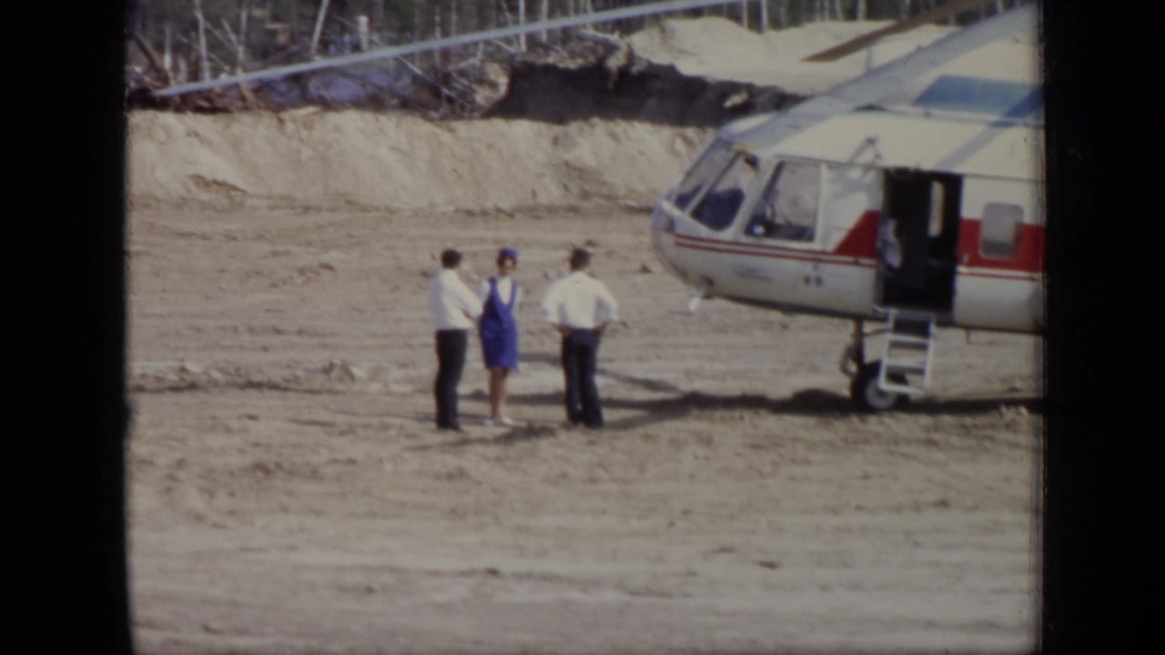 Three adults stand beside an open helicopter on a dirt helipad.
