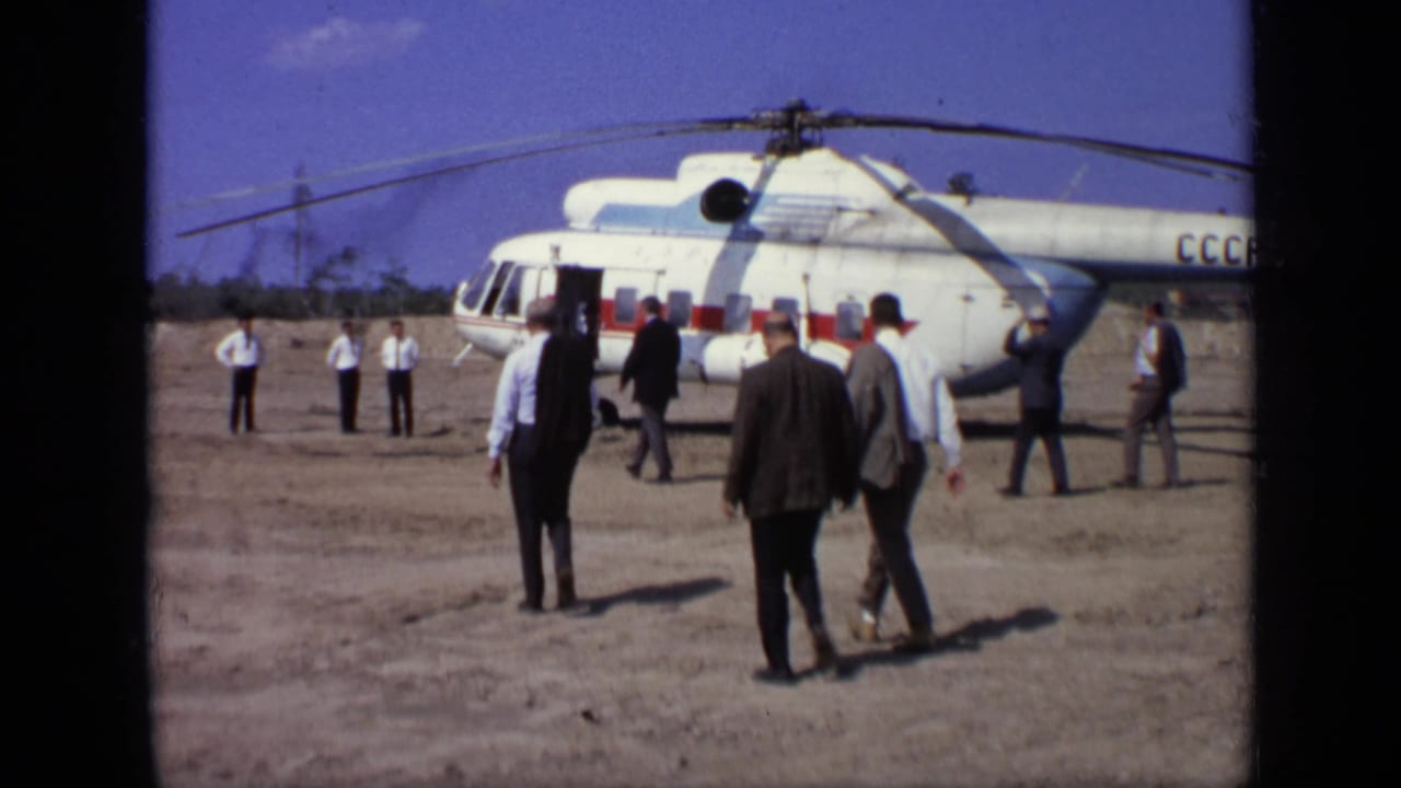 Men walk across a dirt airfield toward a white and red helicopter.