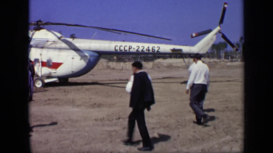 Two men pass the rear of the CCCP-22462 helicopter, with tail rotor, fuselage marking, and open dirt field visible.