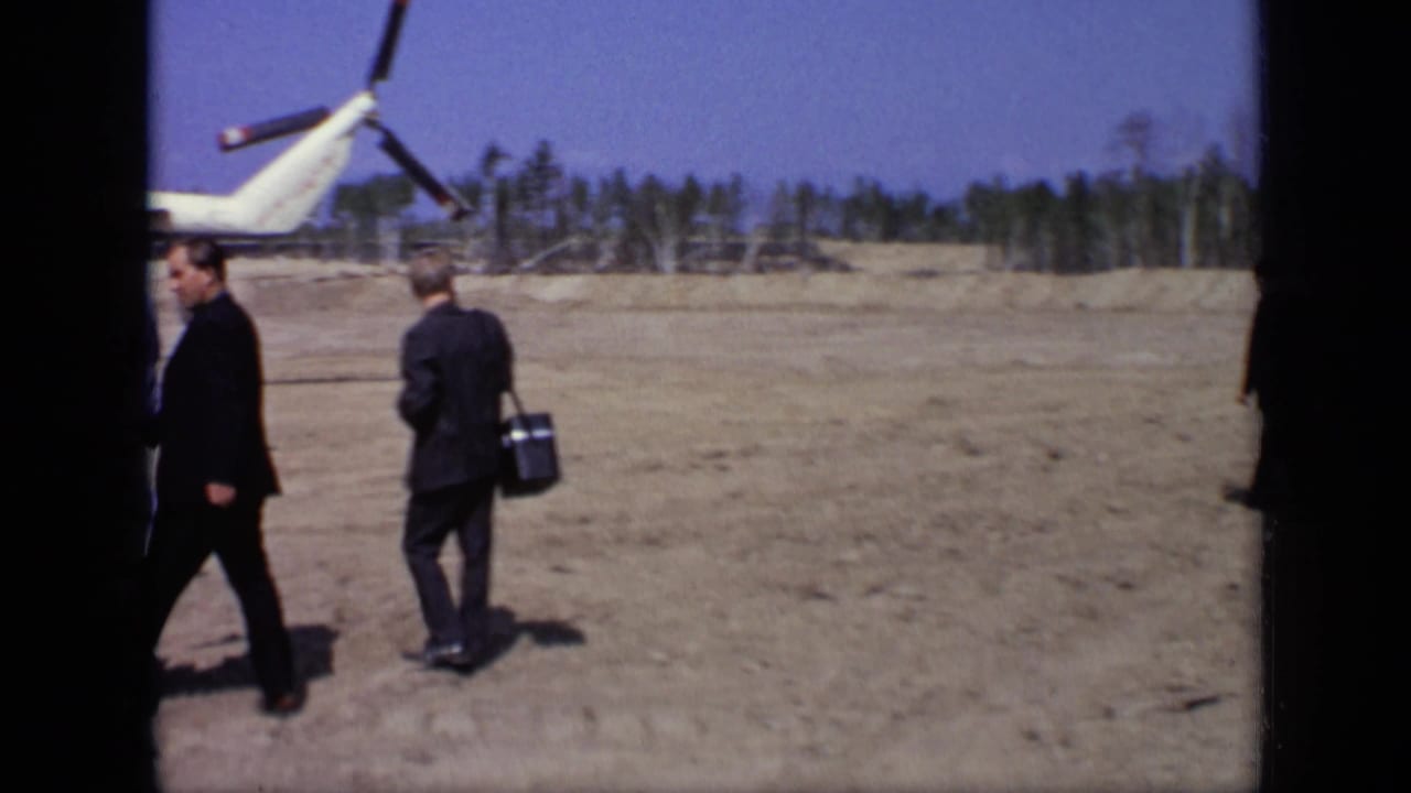 Two suited men walk past the tail area of a white helicopter on open ground.