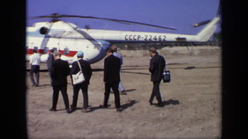 The group stands and walks near the white Soviet helicopter marked CCCP-22462, framed by dry ground, blue sky, and a distant tree line.