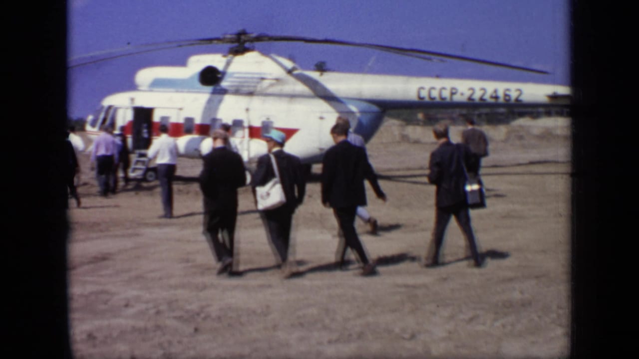 In Moscow, 1971, men in suits and hats walk toward a white-and-red Soviet helicopter marked CCCP-22462 on a dusty airfield.