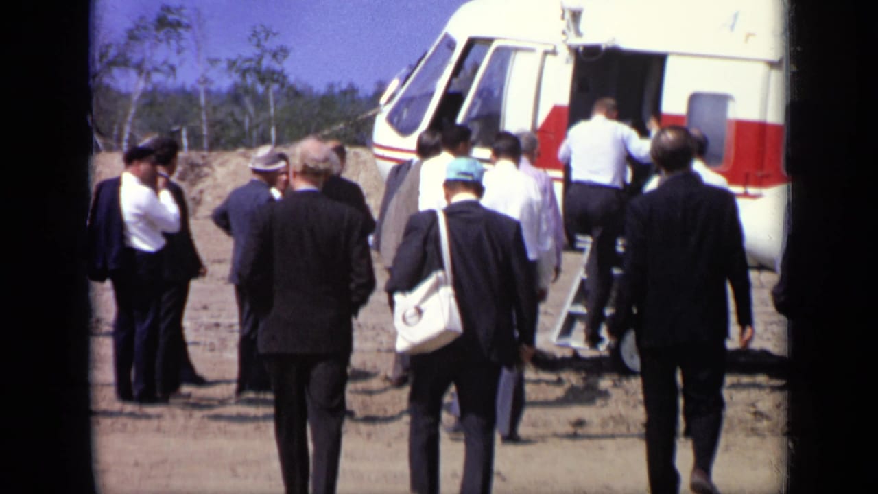 Adults in suits approach a red-and-white helicopter on a dirt airfield.