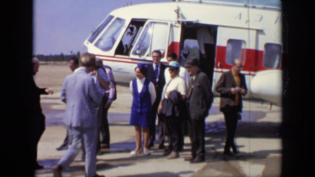 1971 Moscow airfield scene with adults in suits and a woman in blue standing near a white and red helicopter.