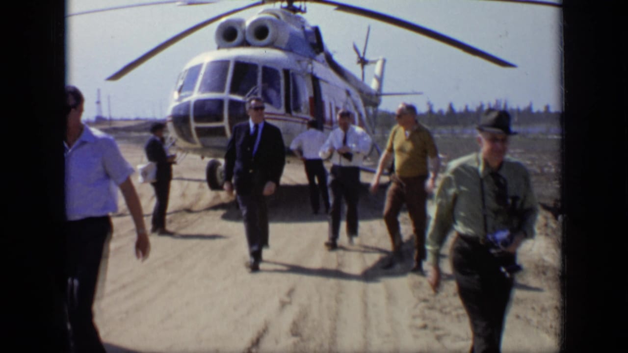 1971 Moscow 8mm frame showing several adult men leaving a parked blue and white helicopter on a sunlit dirt airfield.