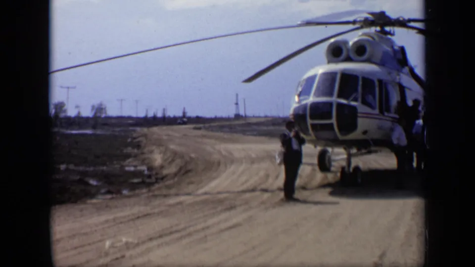 1971 8mm frame from Moscow, Russia metadata showing a helicopter on a dirt road in a sparse, tree-lined field area.