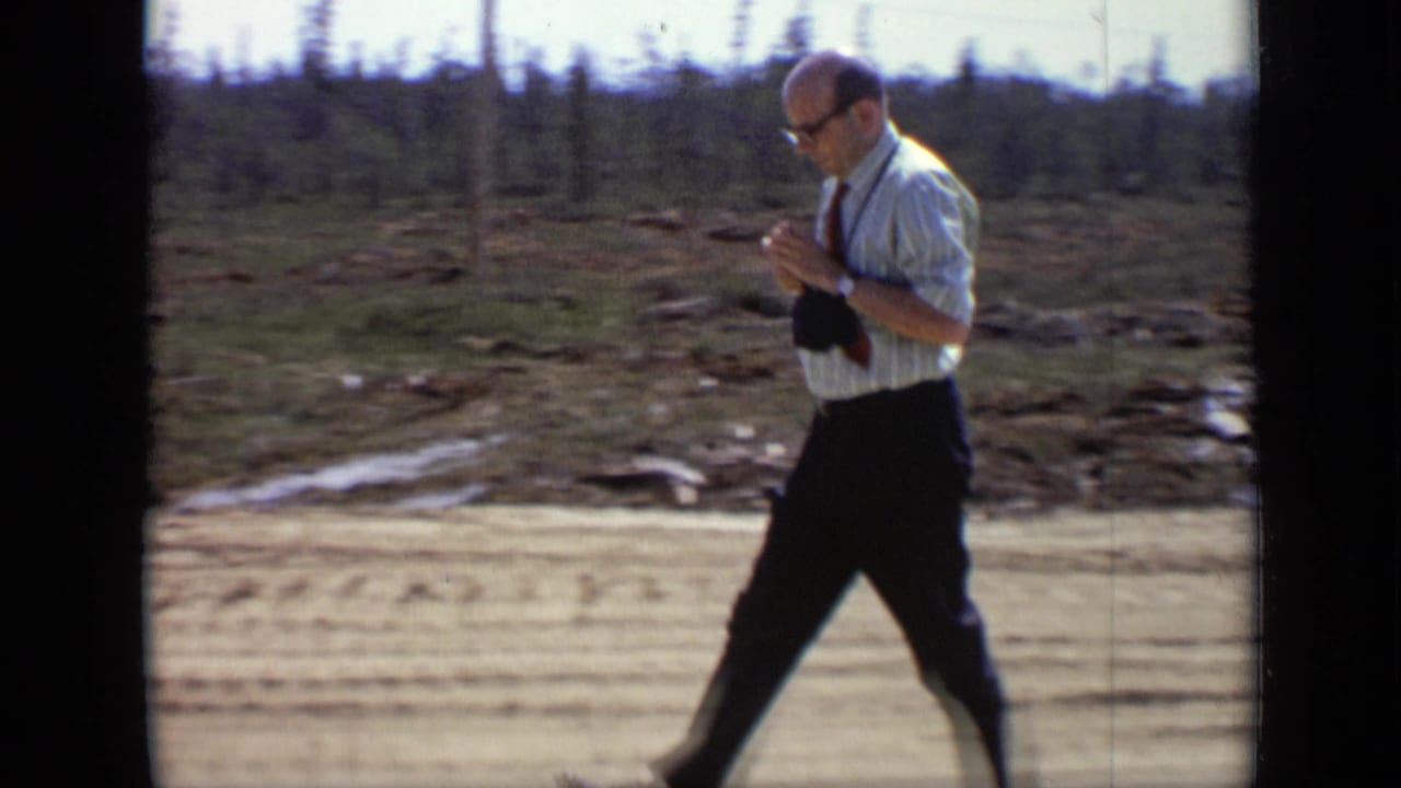 Adult man in glasses and a striped shirt walks beside a remote dirt road, holding equipment in a 1971 8mm field scene.