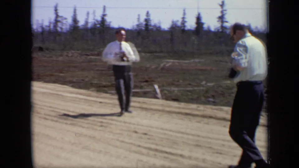 Two adult men appear near a dirt road at the field location, one facing the camera and one partly turned away, in 1971 8mm footage.