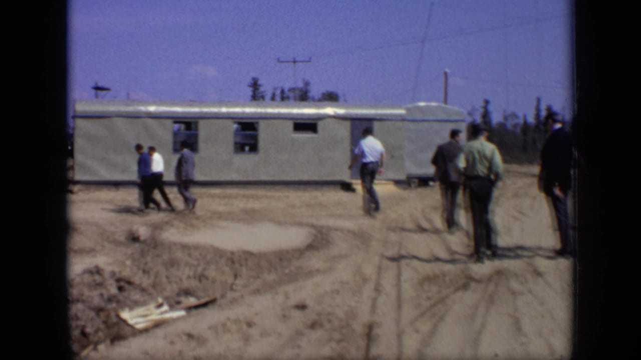 In 1971 Moscow footage, men in dress shirts and suits walk near a long prefabricated building on a dusty construction site.