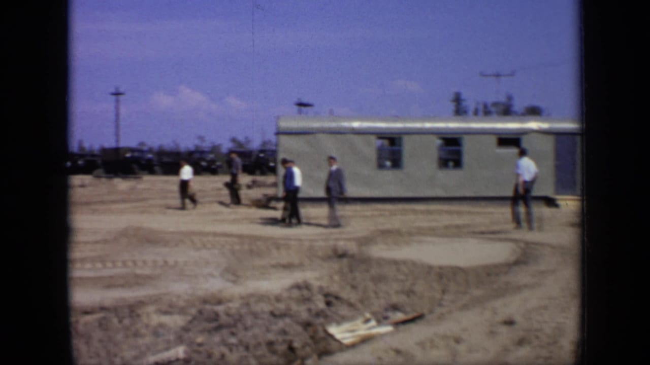 Men in business clothing move across an open Moscow lot with a prefabricated building, cars, and utility poles in the background.