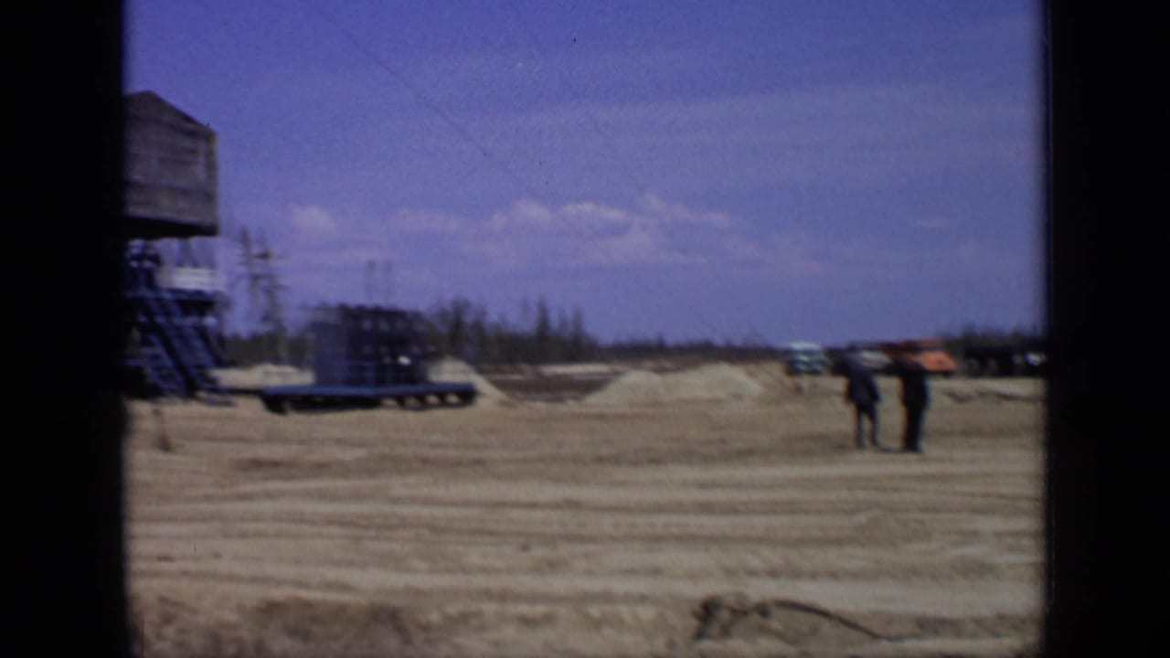 Remote industrial site with workers, flatbed trailers, and a raised structure at the edge of frame.