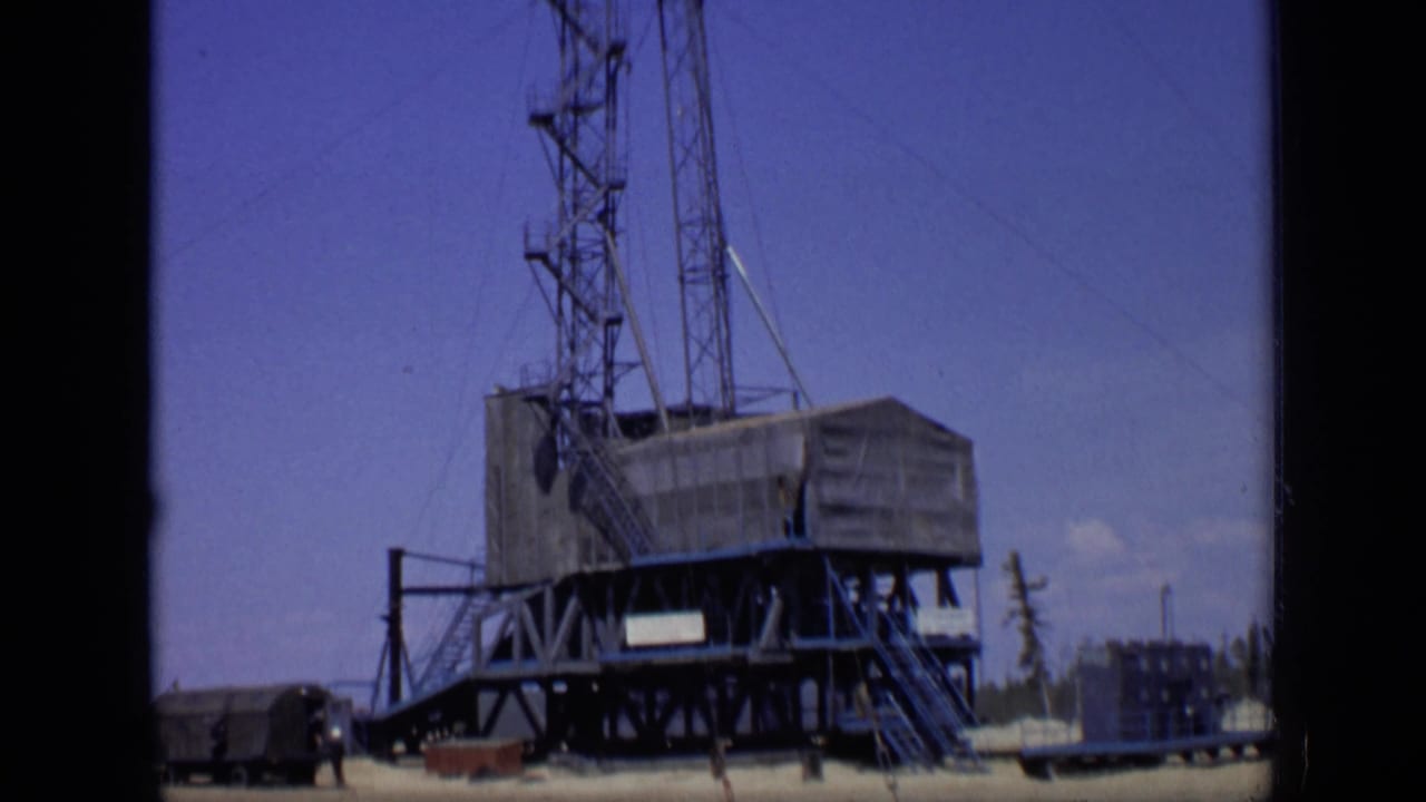 Wide view of a drilling rig base and derrick under a blue sky.