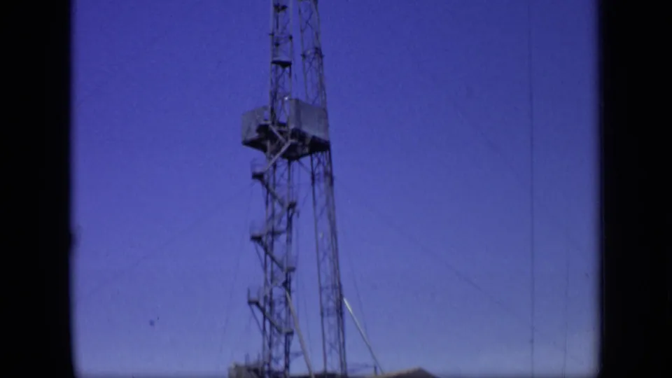 A closer upward view isolates the drilling derrick, showing lattice sections, guy wires and a raised platform in the 8mm frame.
