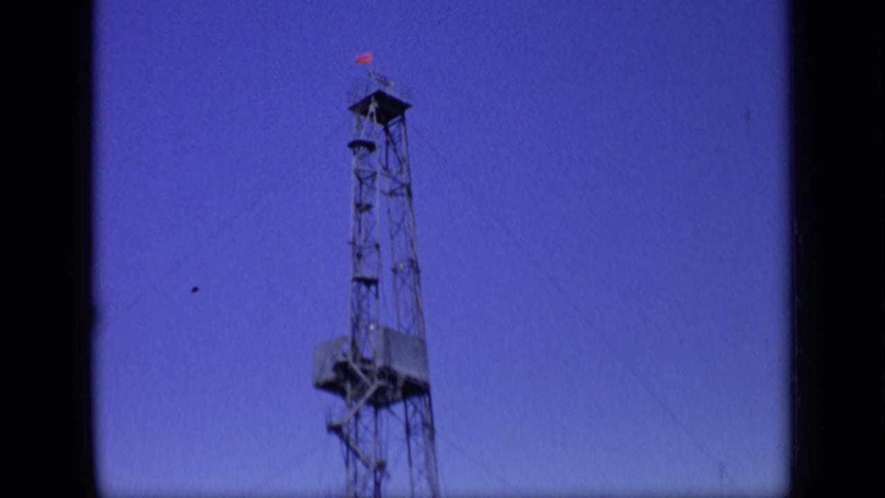 Tall metal drilling rig with a small red flag against a deep blue-purple sky.
