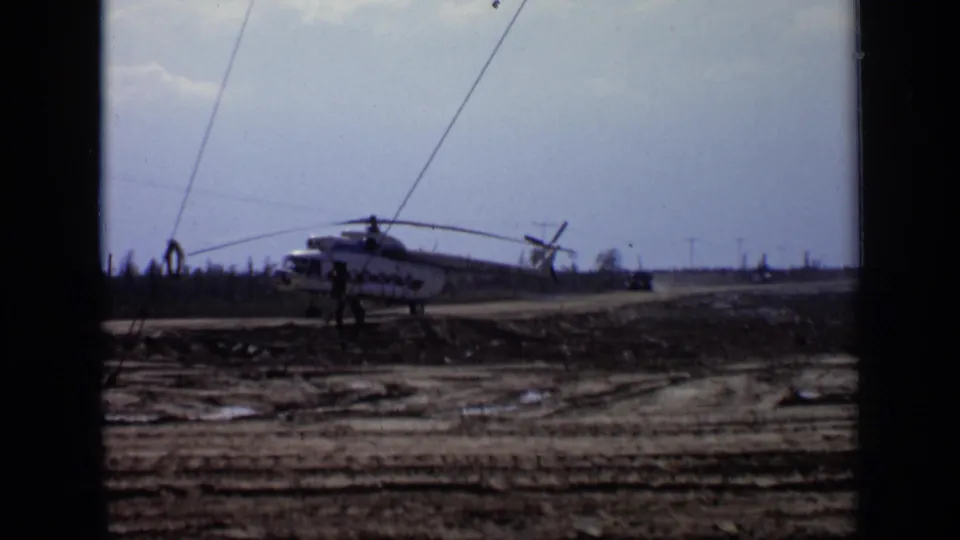1971 Moscow 8mm frame showing a grounded helicopter on muddy open ground, with small distant figures nearby under a pale sky.