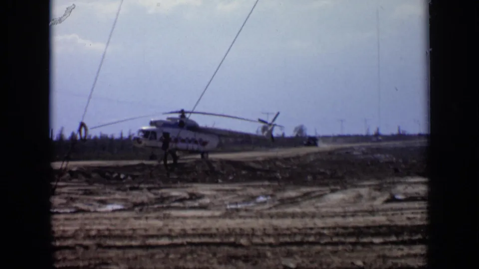 1971 Moscow 8mm frame showing a grounded helicopter in a muddy field, distant people, and a low tree-lined horizon.