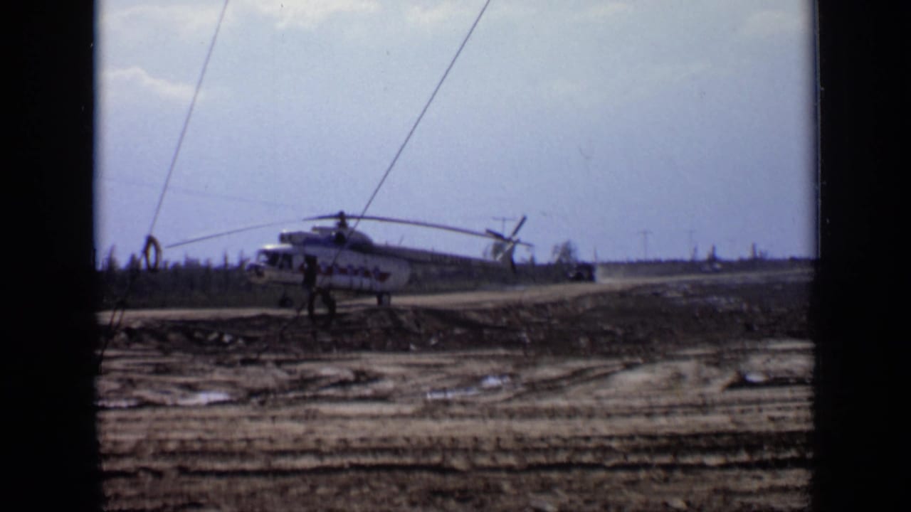 1971 Moscow-area 8mm frame showing a white helicopter with red markings on a muddy forest road as a person stands nearby.