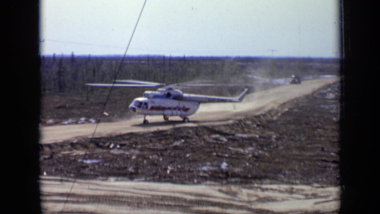 1971 Moscow-area 8mm frame showing a large white helicopter with red markings on a dusty road amid low forest.