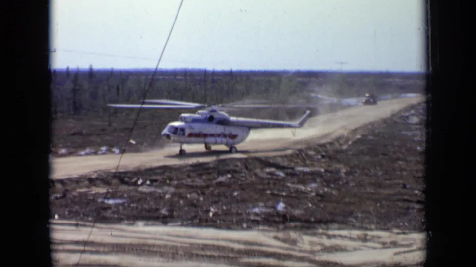 1971 Moscow-area 8mm frame showing a large white helicopter with red markings on a dusty road amid low forest.