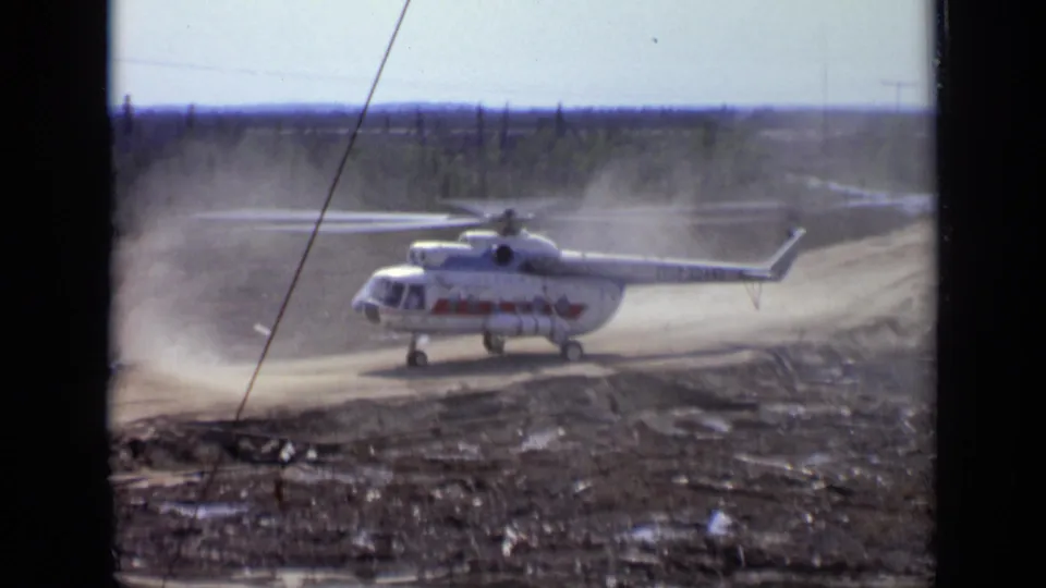 Large white helicopter with red side lettering seen in 1971 Moscow-area 8mm footage, surrounded by dust on a dirt road.
