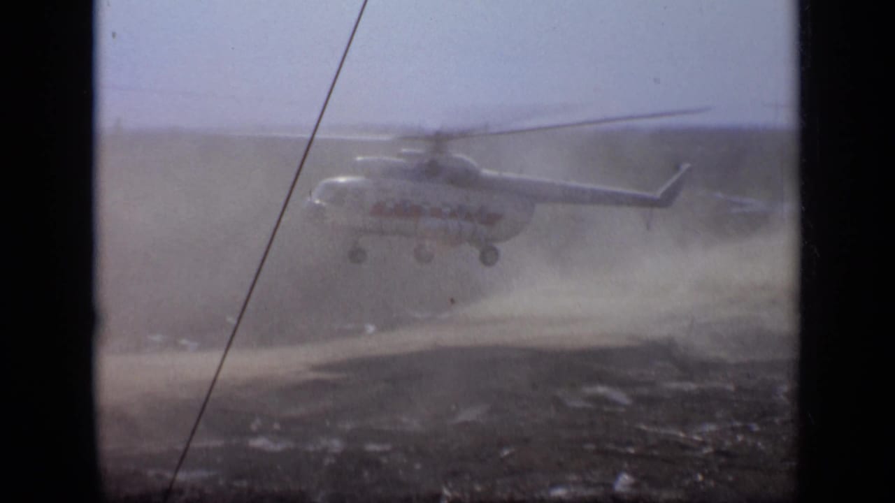 1971 Moscow 8mm frame showing a helicopter lifting low over a dusty open field, seen through soft grain and dark film gate edges.