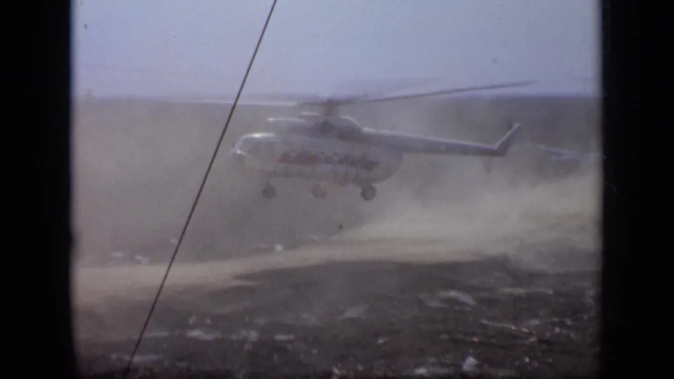 1971 Moscow 8mm frame showing a helicopter lifting low over a dusty open field, seen through soft grain and dark film gate edges.