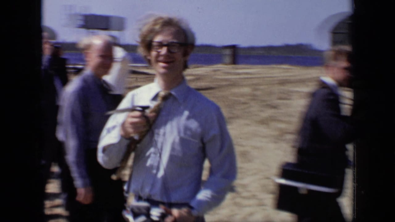 Men at a construction site, with a young man holding a microphone and smiling toward the camera.