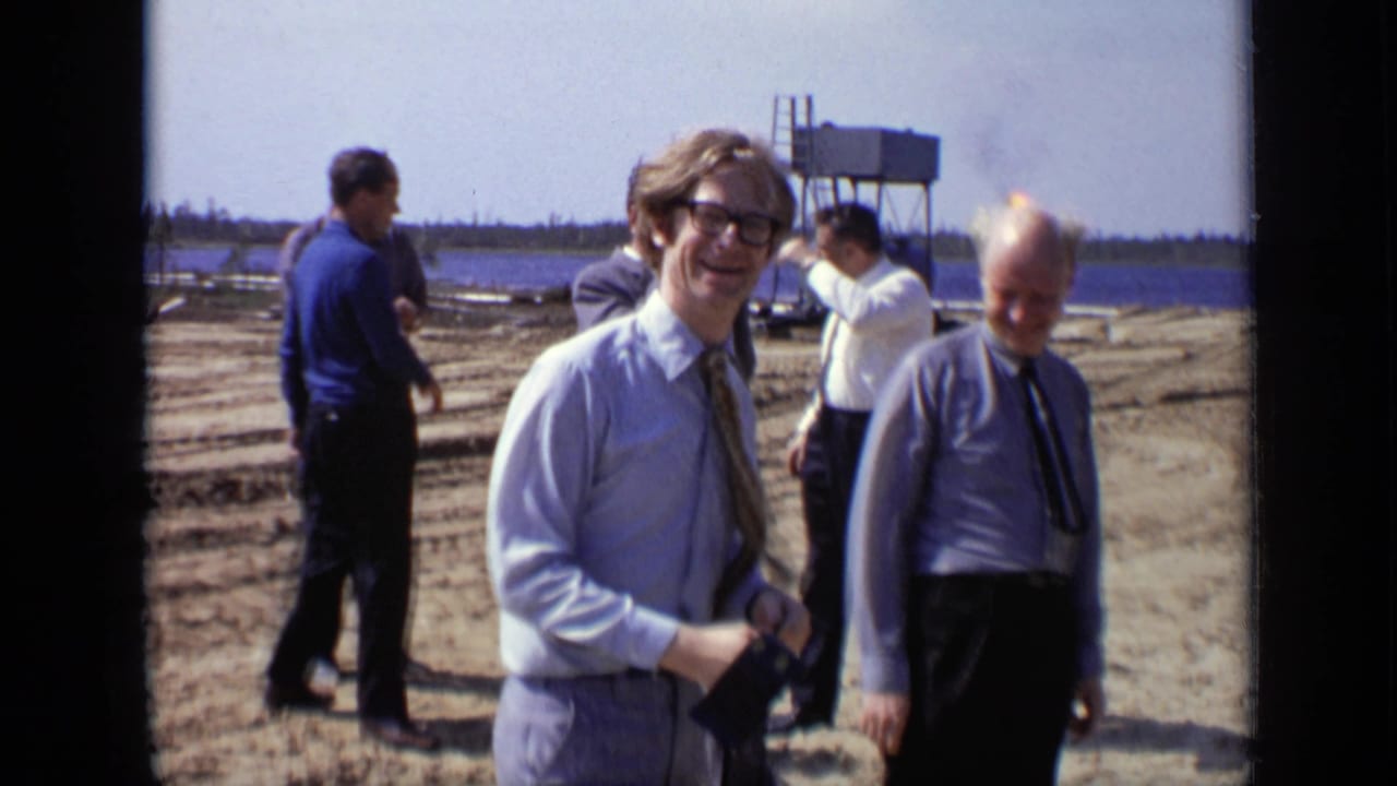 1971 Moscow construction-site view with several men near a waterfront, a microphone in hand, and a distant utility structure behind them.