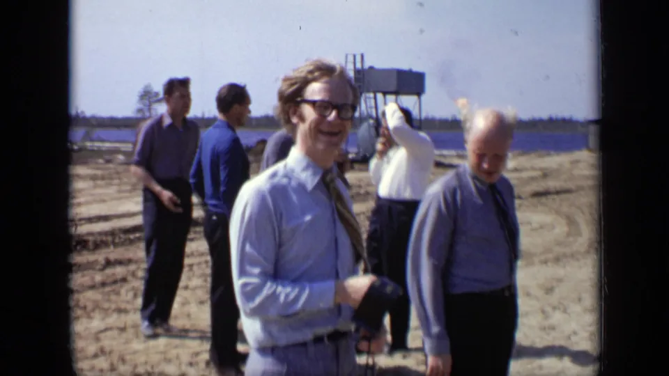 1971 Moscow 8mm frame of men at a windy outdoor construction site, with a smiling microphone holder and others looking around.
